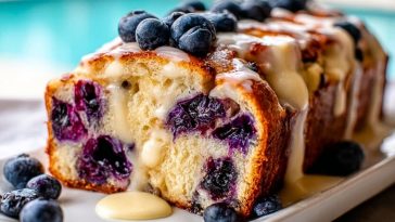 Golden Blueberry Cream Cheese Bread loaf sliced open with creamy glaze dripping, topped with fresh blueberries, beside a pool.