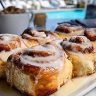 Cinnamon Rolls on a white tray by the pool, dripping with vanilla glaze and showing golden flaky layers.