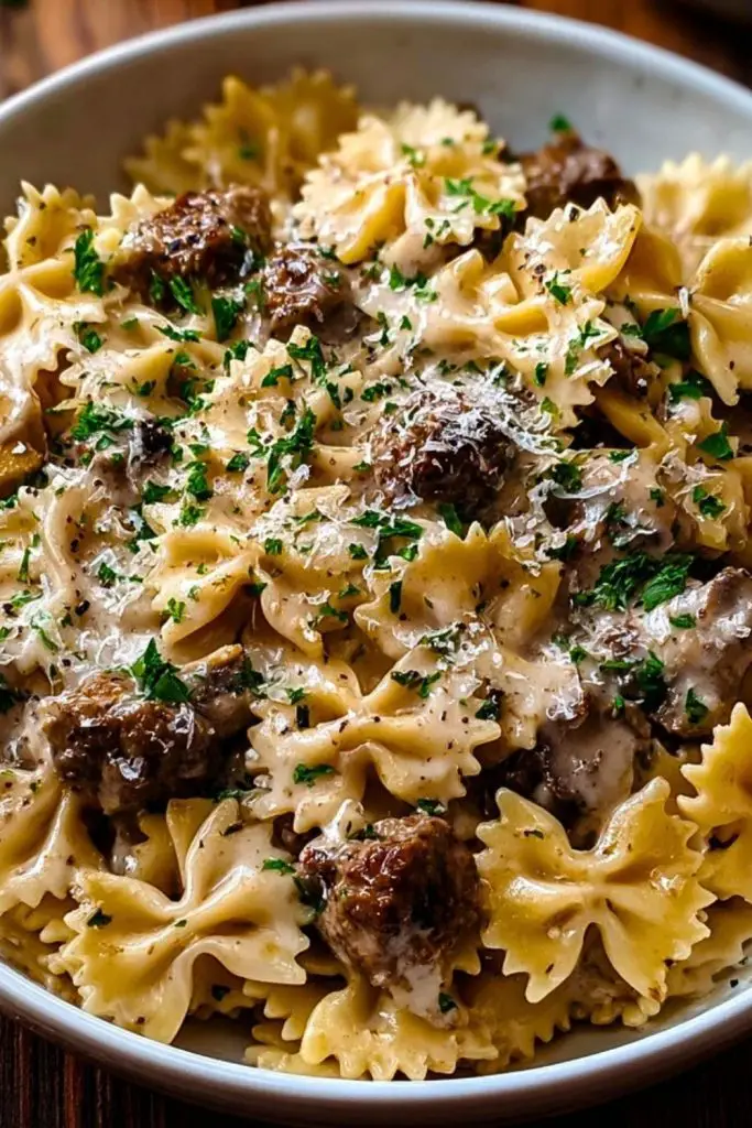 Overhead view of Creamy Beef and Bowtie Pasta with grated parmesan, fresh herbs, and bowtie pasta in a wide white bowl on wooden surface.