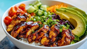 Sliced honey chipotle chicken over white rice with avocado, tomatoes, cucumbers, carrots, and cabbage in a white bowl.