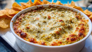 Bubbly Hot Artichoke Dip served in a white baking dish with golden cheese top, surrounded by crispy tortilla chips by the pool.