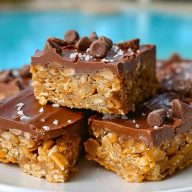 Chewy Special K Bars topped with chocolate chips and flaky sea salt, stacked on a white plate near a pool.
