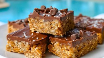 Chewy Special K Bars topped with chocolate chips and flaky sea salt, stacked on a white plate near a pool.