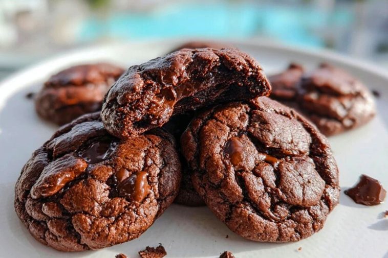 Double Chocolate Cookies stacked on a white plate with melty chocolate chunks and a poolside view