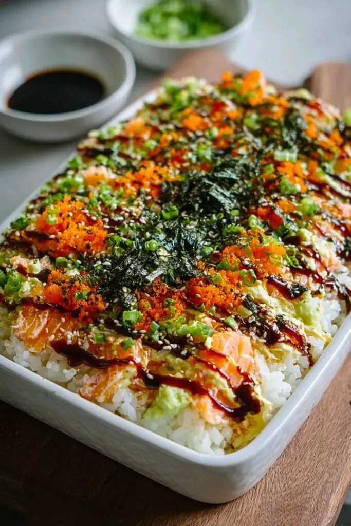 Casserole of Easy Sushi Bake topped with avocado, tobiko, seaweed, green onions, and sauce drizzle, served with soy sauce on the side.