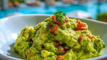 Fresh guacamole served in a white bowl, topped with diced tomato and parsley, with a sunny poolside background.