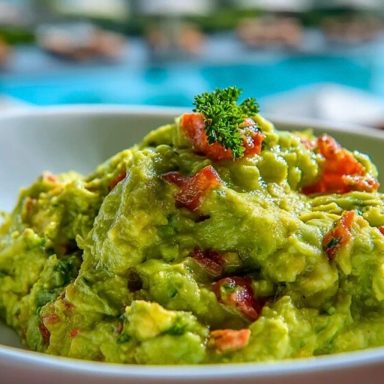 Fresh guacamole served in a white bowl, topped with diced tomato and parsley, with a sunny poolside background.