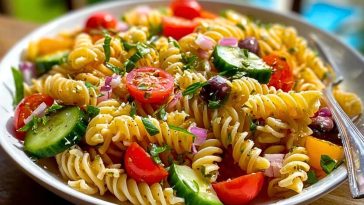A bowl of Pasta Salad with Italian Dressing featuring rotini pasta, cherry tomatoes, cucumbers, black olives, and feta, garnished with fresh herbs.