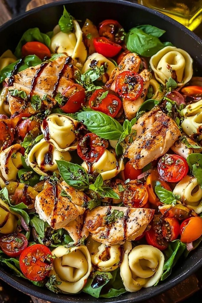 Close-up of Warm Balsamic Chicken Tortellini Salad showing seared chicken, pasta, and tomatoes with herbs and balsamic glaze over a bed of greens.